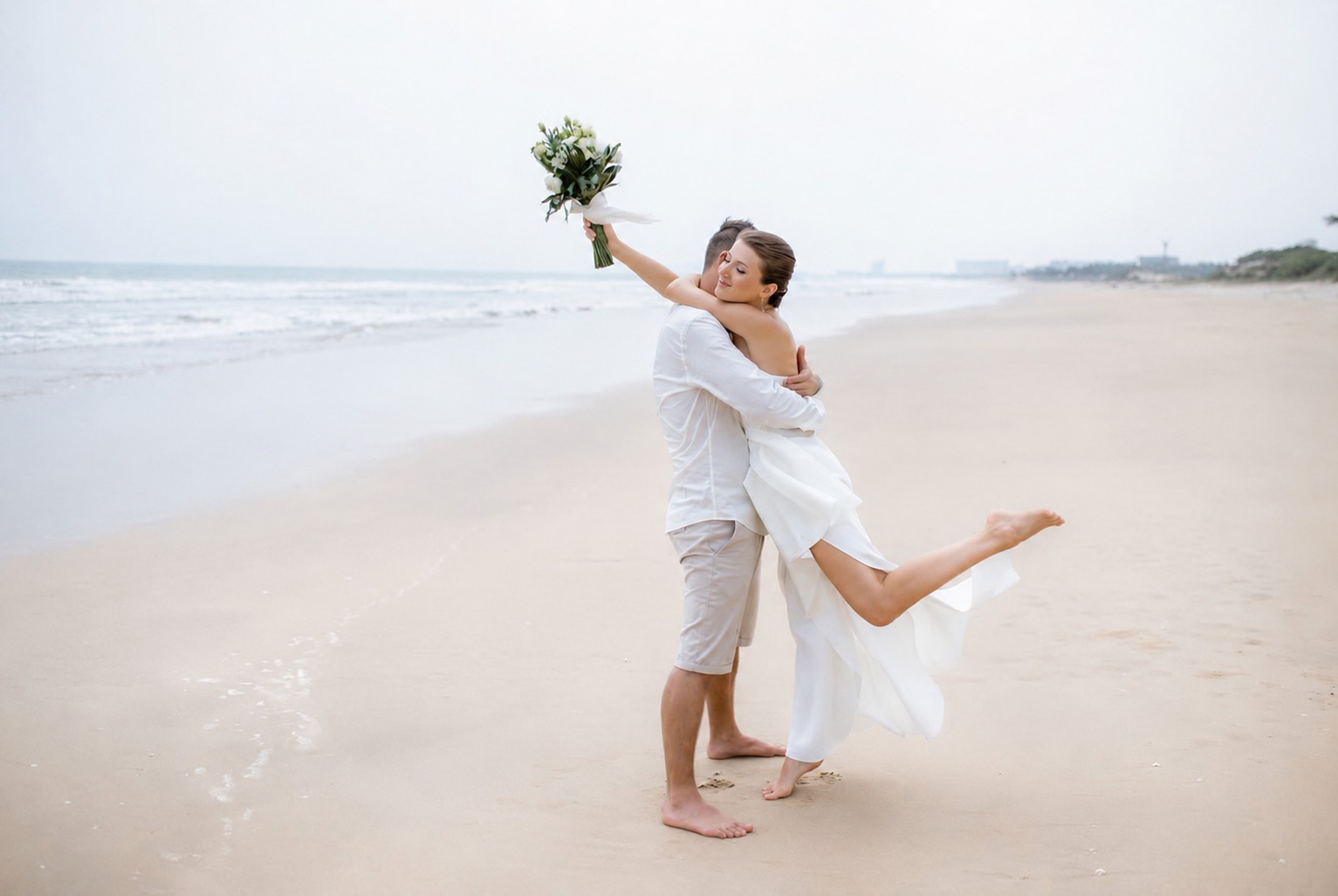 Couple embracing barefoot on an empty beach, bride holding her bouquet aloft