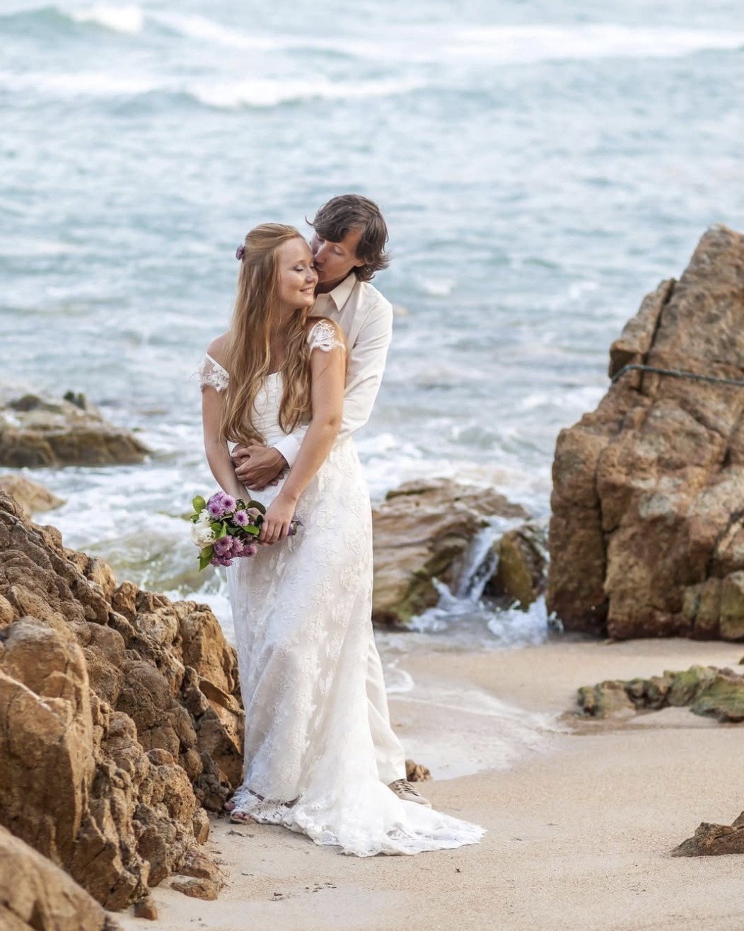 Bride in a lace gown with a bouquet of pink blooms as the groom kisses her temple, on the sand between rocks at the water’s edge