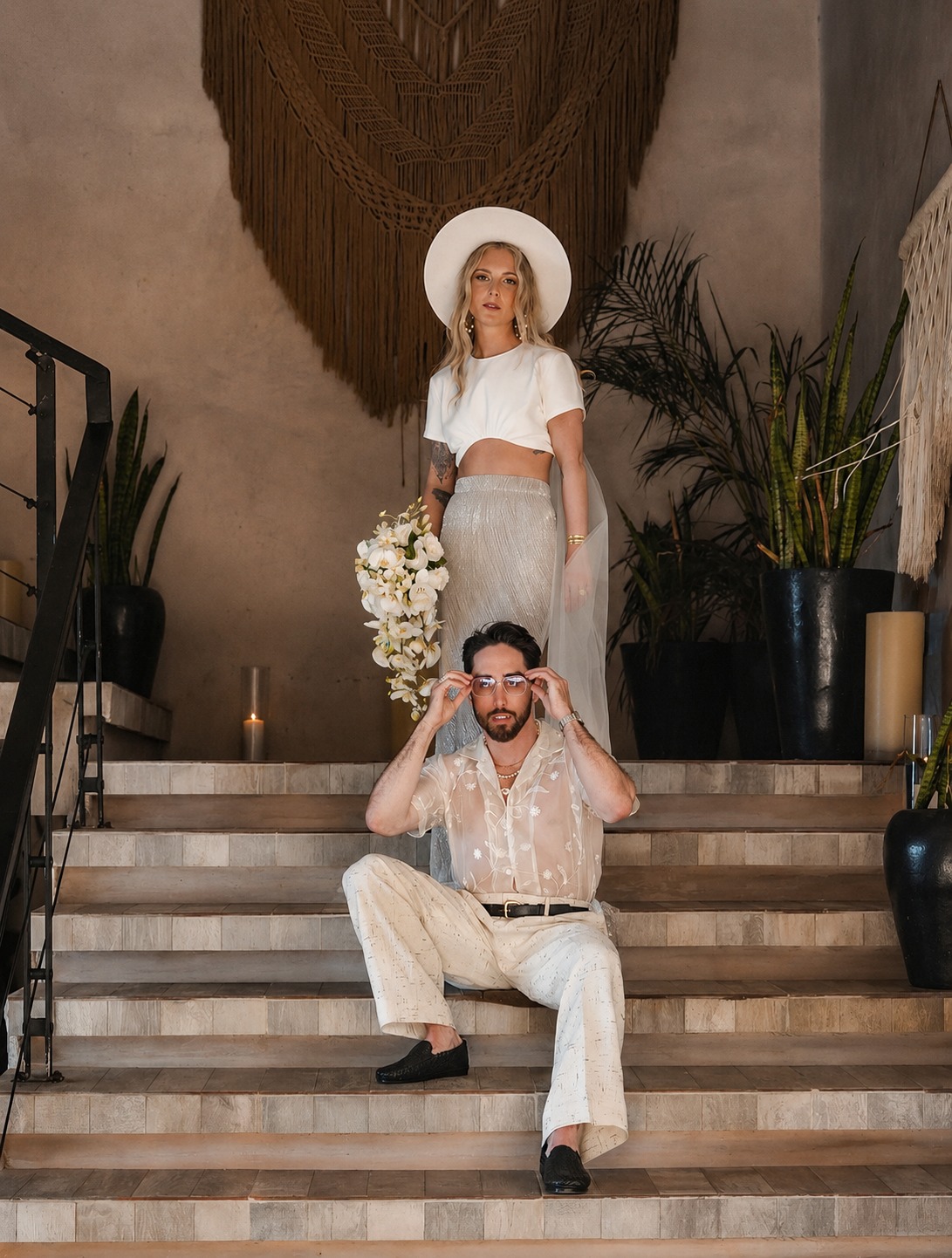 Editorial couple session on a stone staircase: she stands at the top in a wide-brimmed hat holding white orchids, he sits on the steps adjusting his sunglasses, macramé and plants behind them