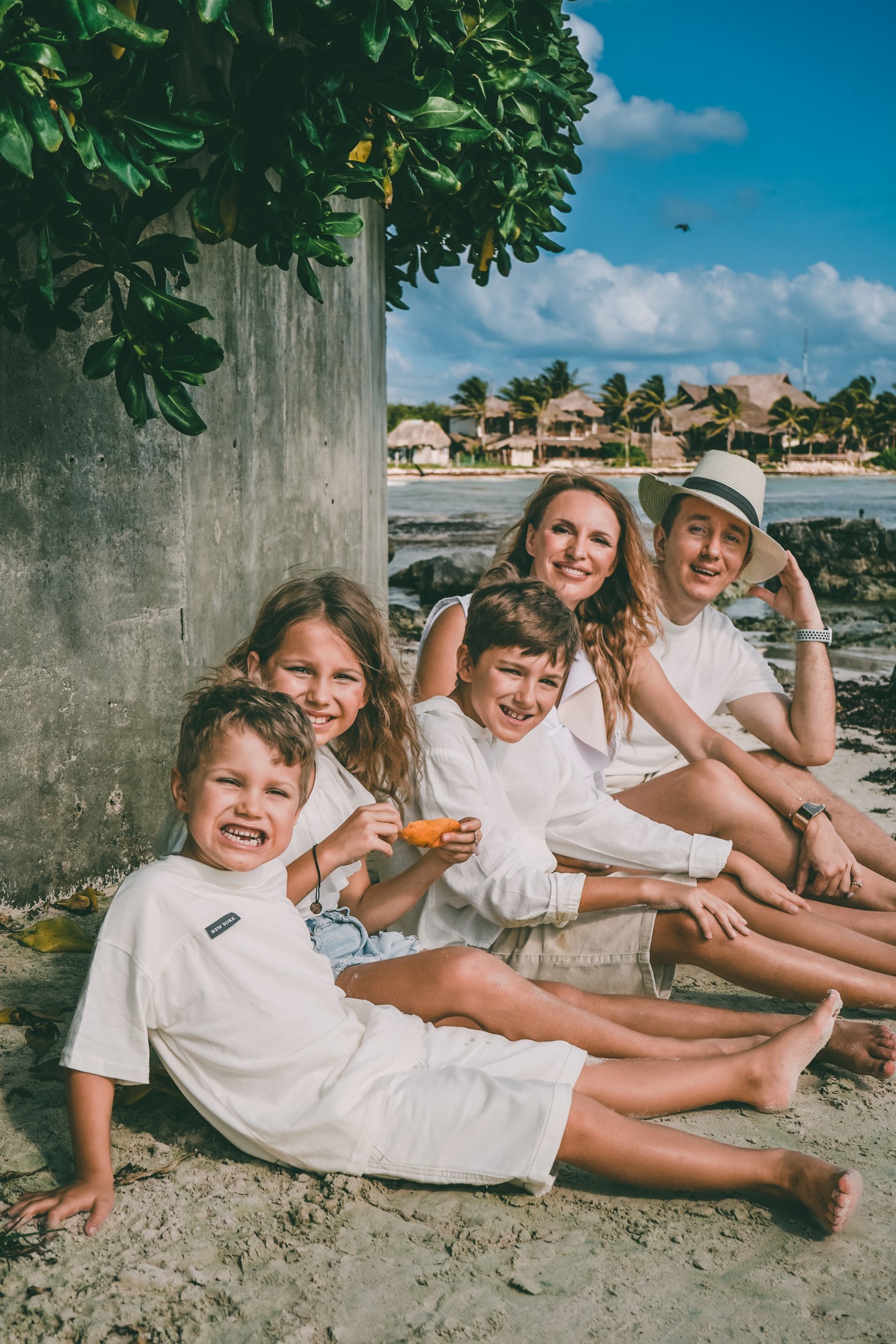 Family of five in white linen sitting together on the sand, laughing, with thatched palapas and the sea behind them