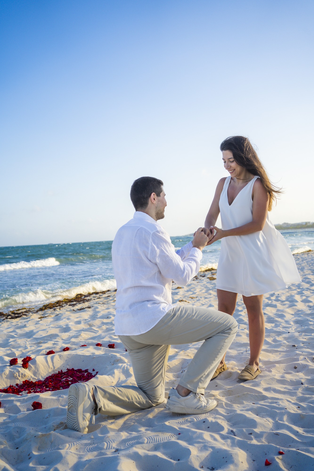 Marriage proposal on the sand: he kneels and takes her hand beside a heart of red rose petals, Riviera Maya